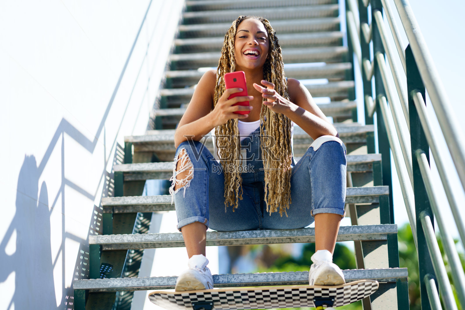 一个黑人女孩,梳着彩色的辫子,坐在那里拿着智能手机和滑板。照片摄影图片