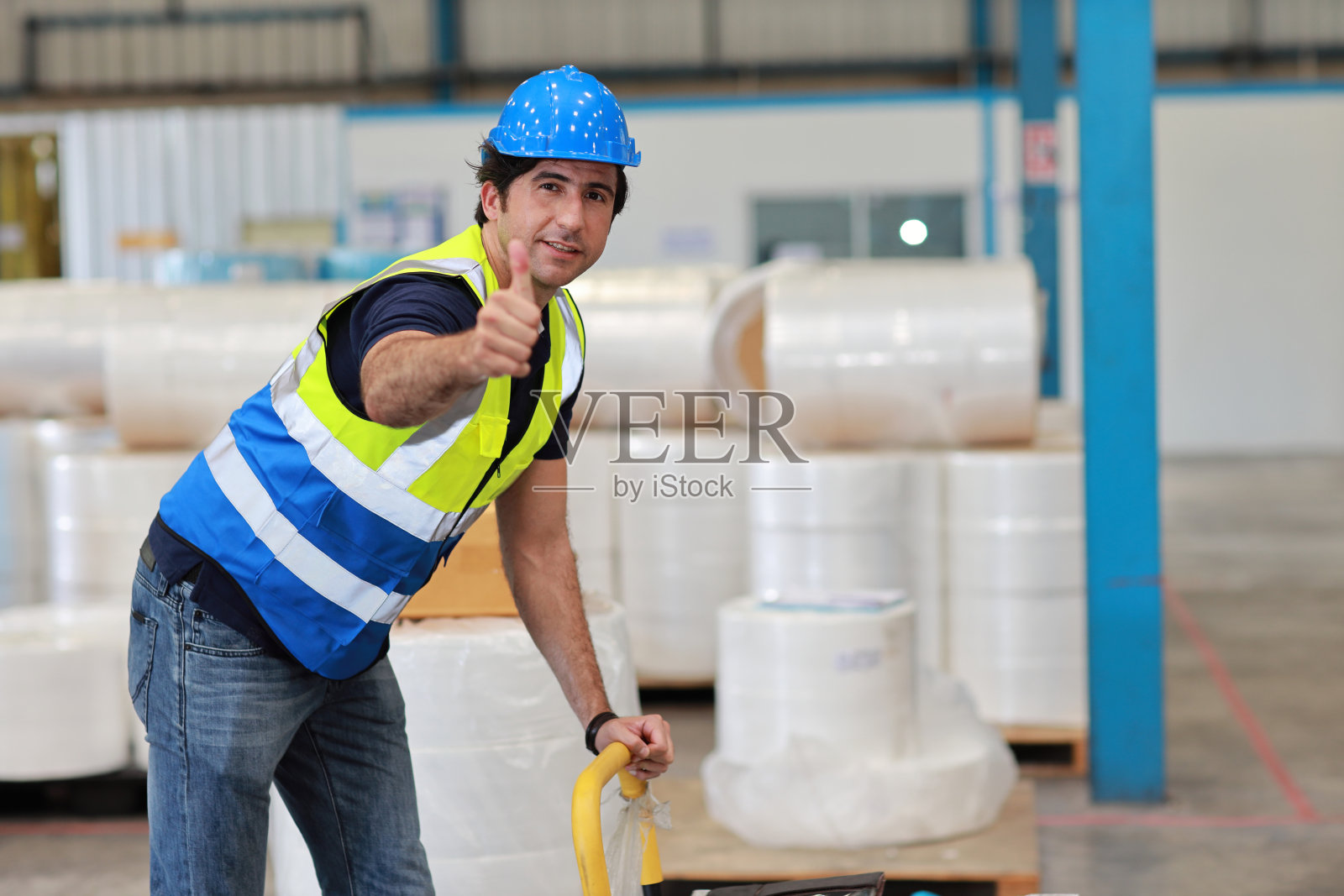 仓库工人在大型仓库中拉托盘车,将包装箱搬运到货架上。活跃的白人男子露出大腹,微笑着看着镜头。照片摄影图片