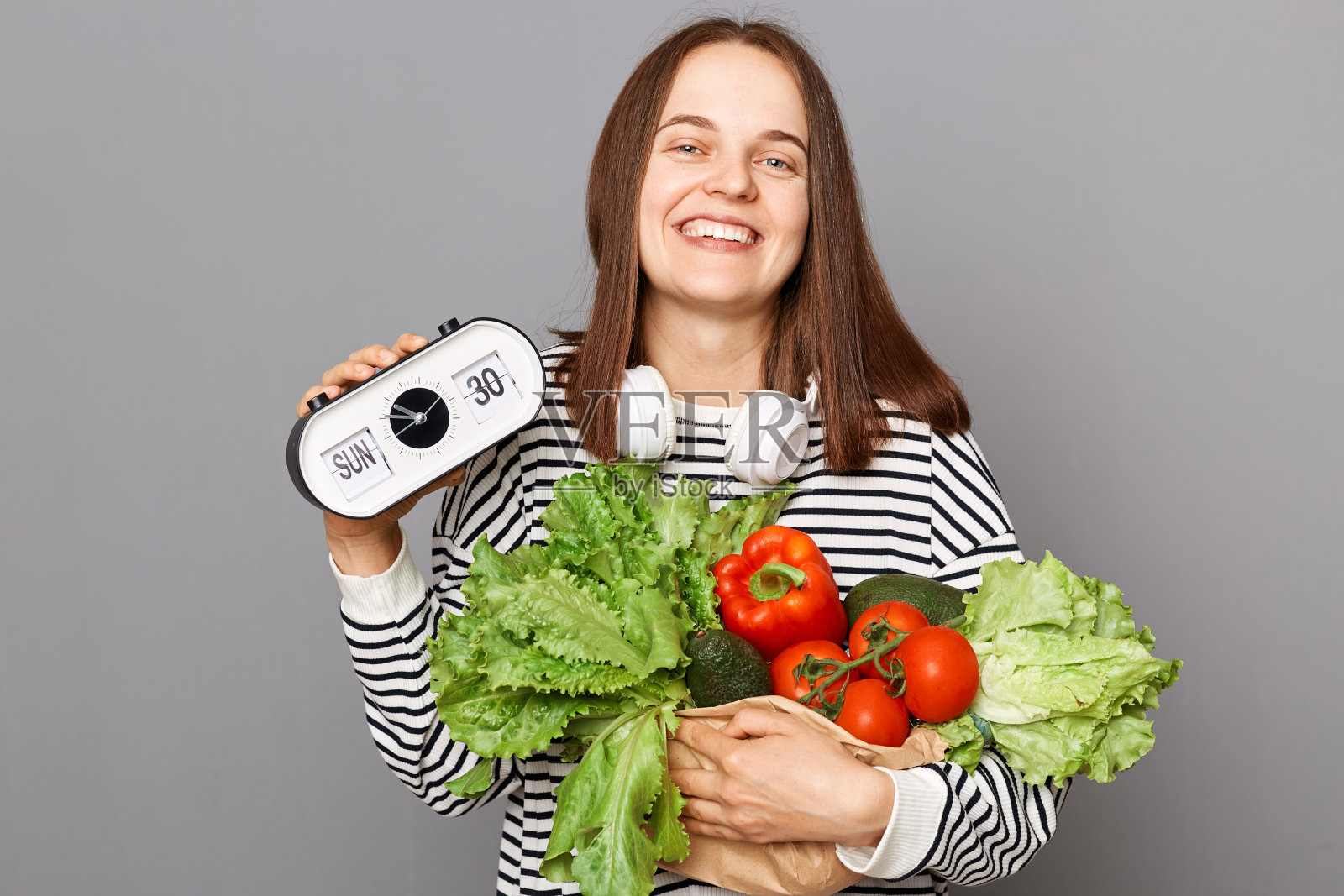 是时候吃新鲜食物了。素食促进健康。健康的生活方式,绿色的选择。微笑的女人拿着蔬菜,展示着灰色背景上孤立的时钟。照片摄影图片