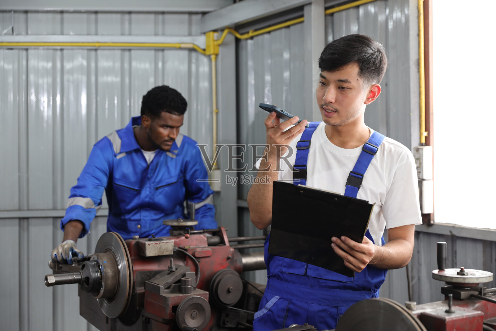 在车库,多民族技师技师使用金属车床用智能手机和剪贴板操作抛光汽车盘式制动器。维护汽车和检查汽车零件的概念照片摄影图片