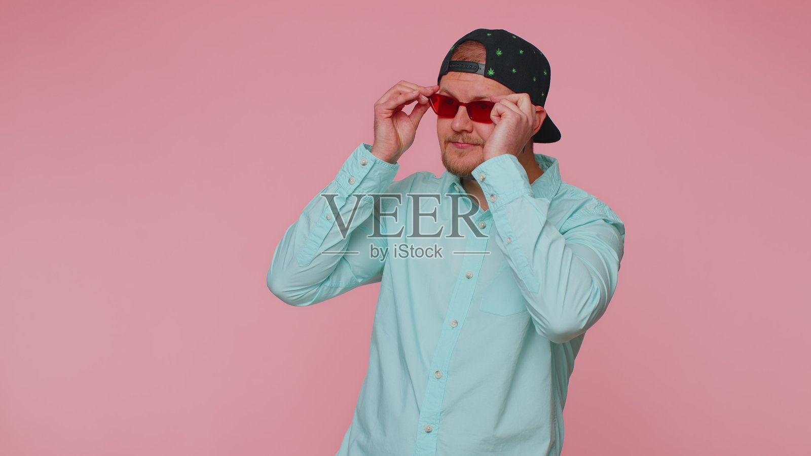 性感开朗的时尚男士穿着蓝色衬衫,戴着墨镜,粉红色的墙上挂着迷人的微笑照片摄影图片