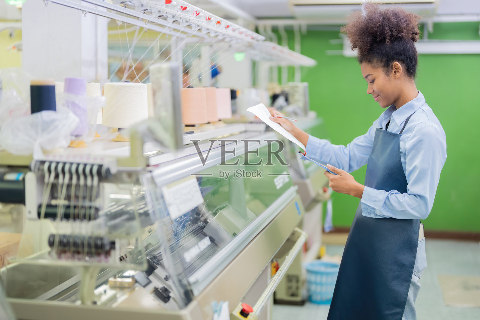 美国女设计师、织女站着展开产品设计订单清单。她旁边有一台正在工作的缝纫机和一卷五颜六色的线。在一个庞大的织造工业中照片摄影图片