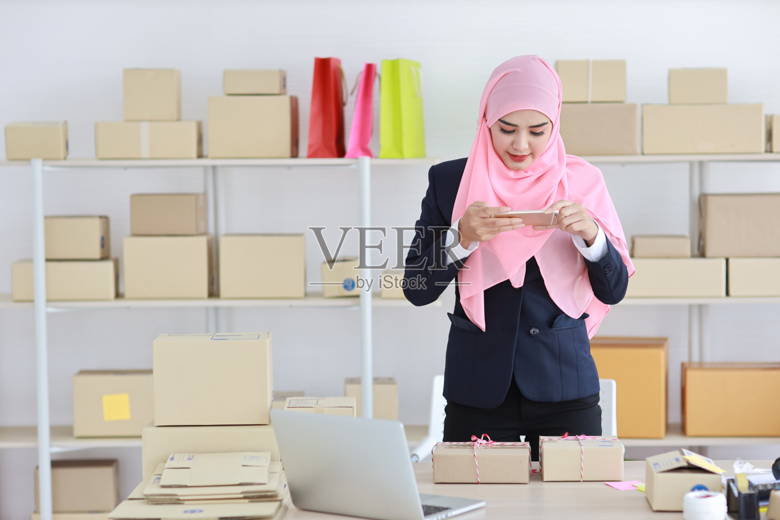 身着蓝色套装的虔诚的亚洲穆斯林妇女站在那里,用手机拍下了送来的包裹。创业中小企业自由职业女孩在家工作,脸上带着幸福的笑容照片摄影图片