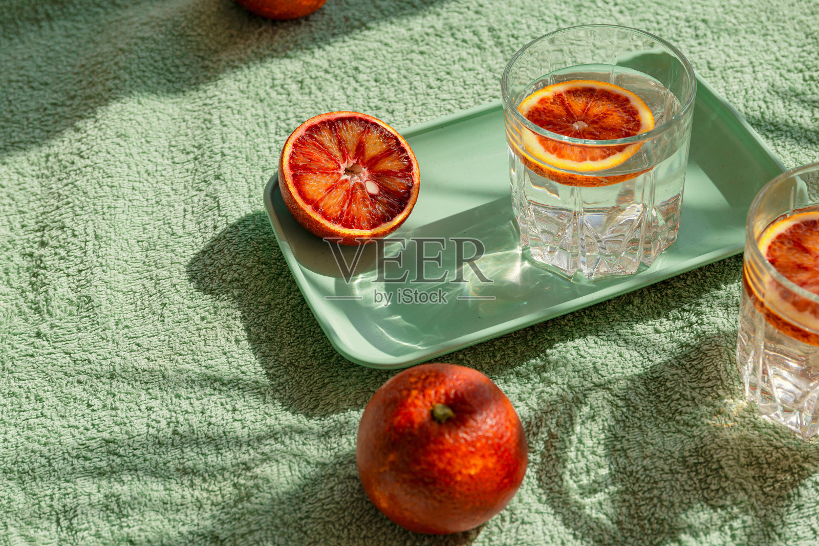 半血橙放在托盘上,两杯鸡尾酒放在绿色沙滩毛巾上,背景是棕榈树树叶的阴影,创意构图。夏日茶点和派对概念。最小的风格。照片摄影图片