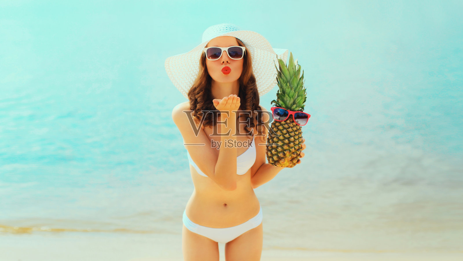 暑假,美丽快乐的年轻女子穿着比基尼泳装,戴着草帽,拿着菠萝水果,在阳光明媚的日子里,在海边背景的海滩上吹着她的嘴唇送吻照片摄影图片