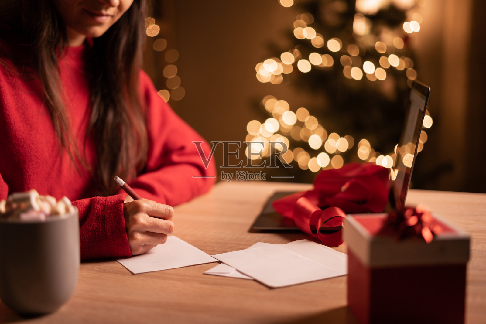 圣诞节期间,穿着红色毛衣的年轻可爱的女人在家里办公室写贺卡或信件照片摄影图片