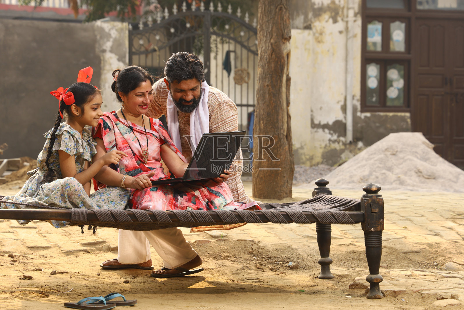 幸福的印度农村家庭。一起坐在自家前院的小床上微笑,用笔记本电脑进行网上支付。妻子穿着漂亮的纱丽。传统的印度文化。农村的生活方式。照片摄影图片