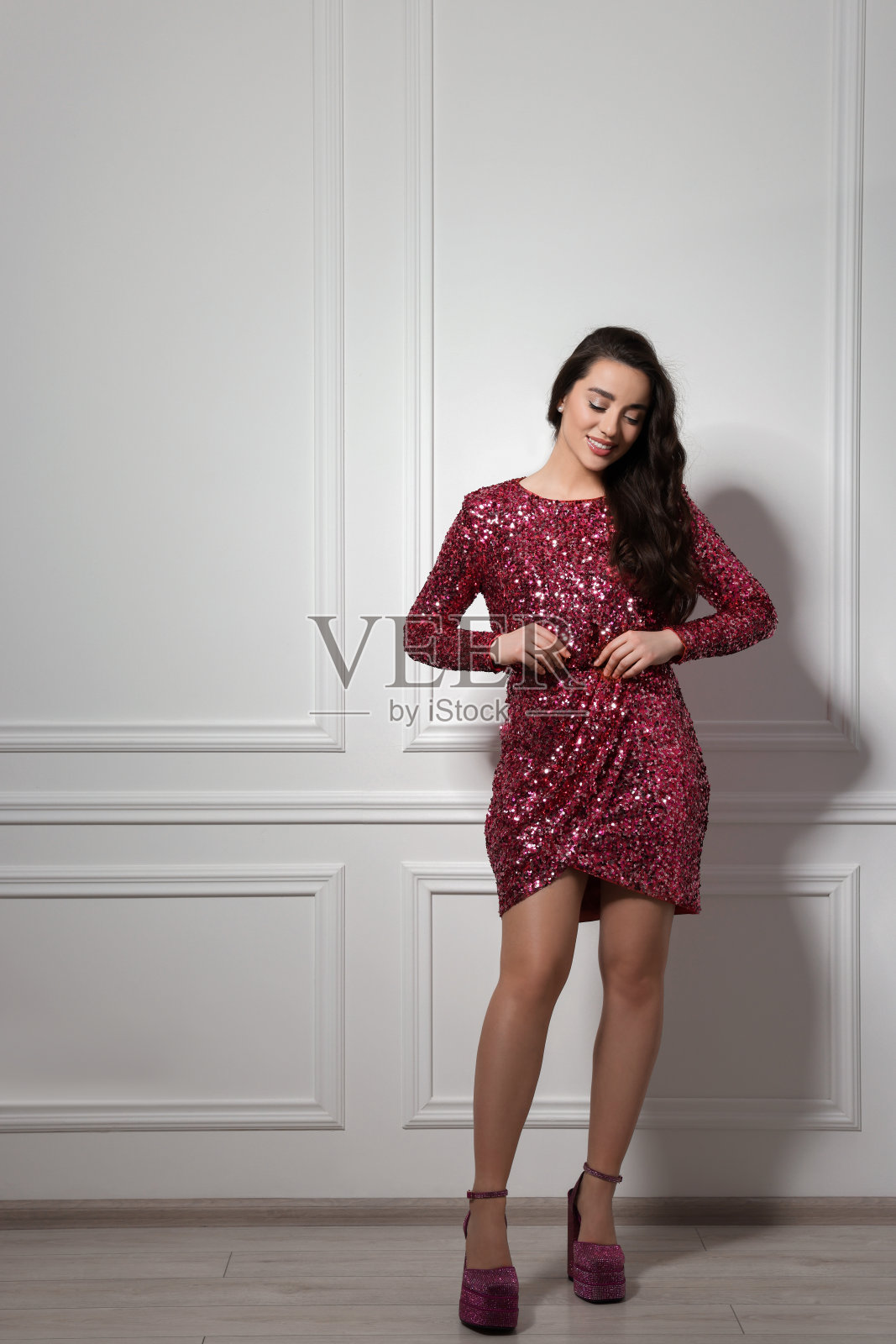 漂亮的年轻女子穿着时髦的粉红色亮片连衣裙,靠近室内的白墙。政派对组织照片摄影图片