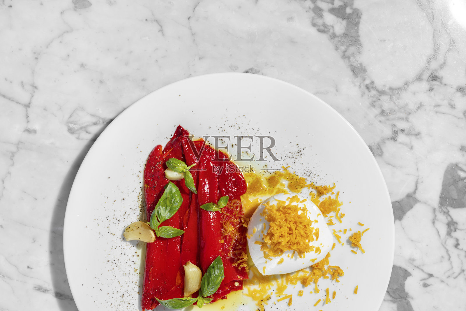 一个浅色的盘子里放着一整团马苏里拉奶酪和薯片,旁边放着一些腌辣椒和罗勒叶蒜。盘子旁边是一杯玫瑰酒。盘子放在一个浅色的大理石台面上。照片摄影图片