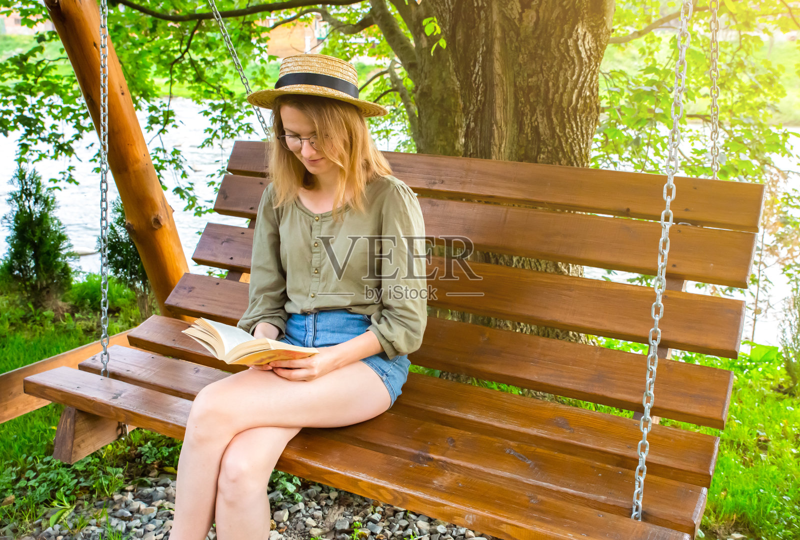 戴眼镜的时髦女孩坐在木凳上看书。女人在户外的门廊上放松,在大自然中荡秋千。舒适的照片。夏天的心情。照片摄影图片