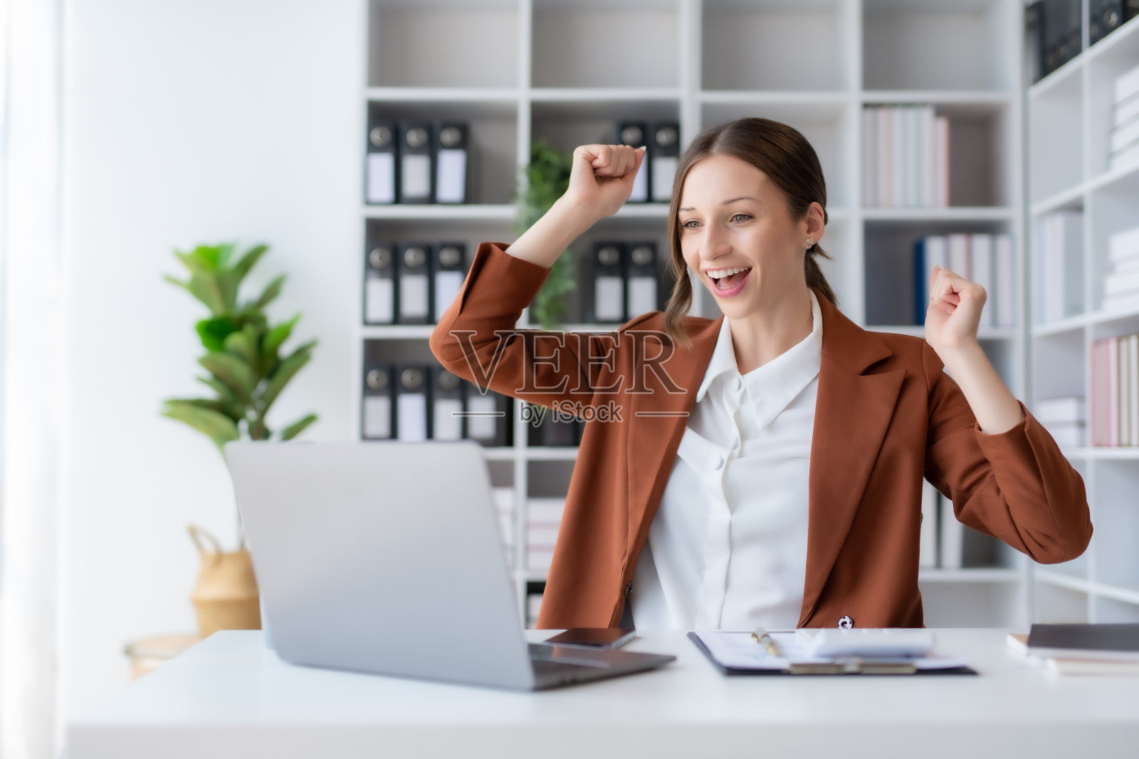 快乐的女商人举起胜利的手,拿着笔记本电脑开心地微笑。工作中成功的概念。照片摄影图片