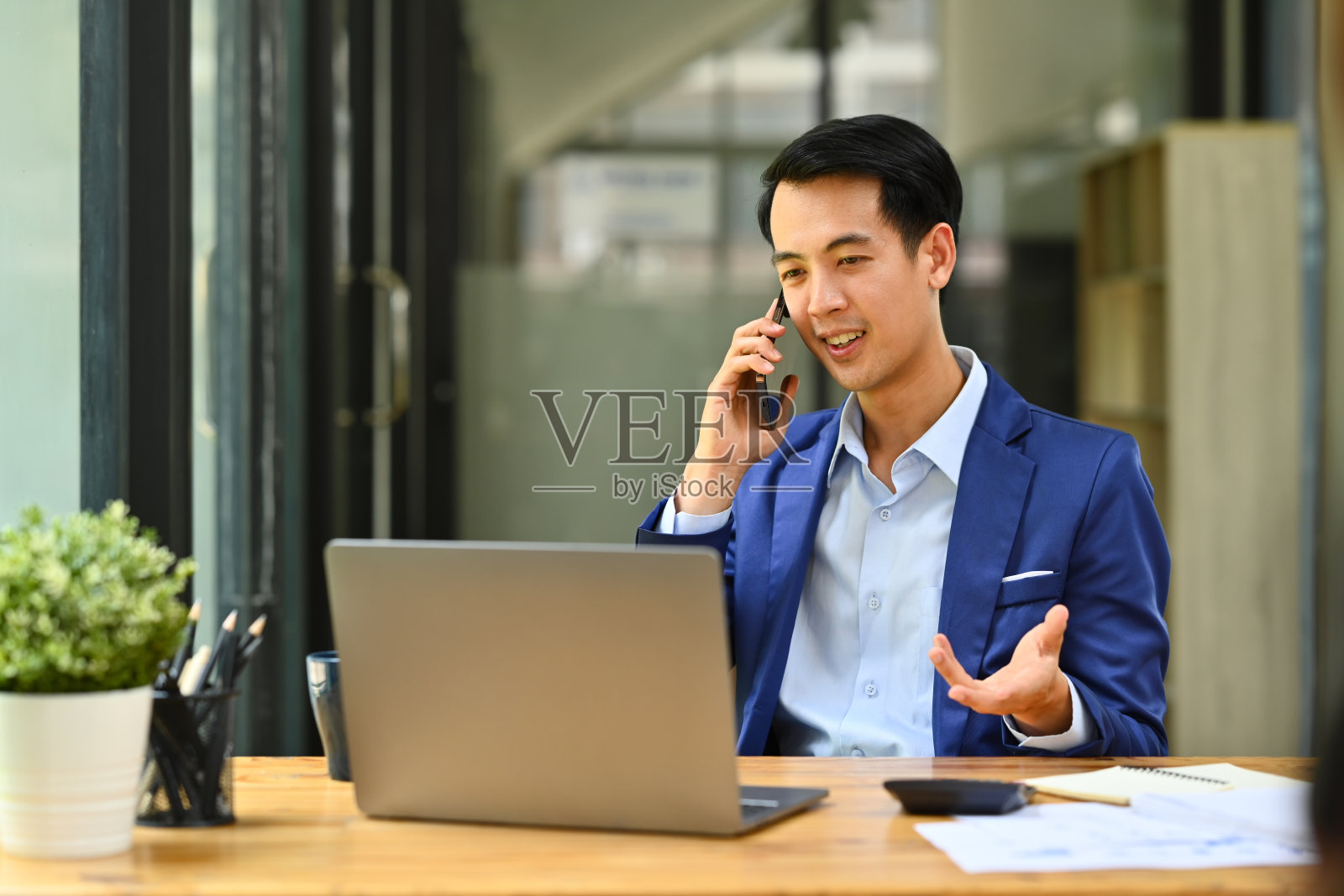 年轻的职业商人穿着漂亮的西装,看着笔记本电脑屏幕,在公司办公室用手机打电话。照片摄影图片