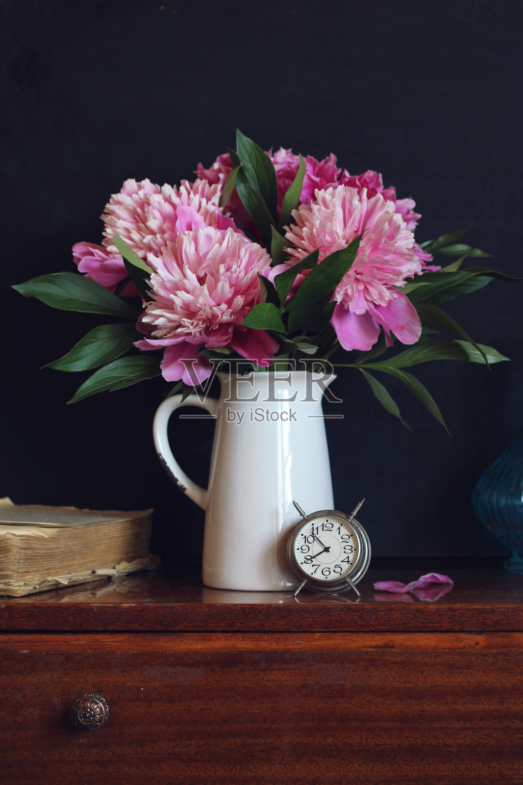 暗色背景上的粉红色牡丹花束。花园里夏天的花,一个钟和一本书。照片摄影图片