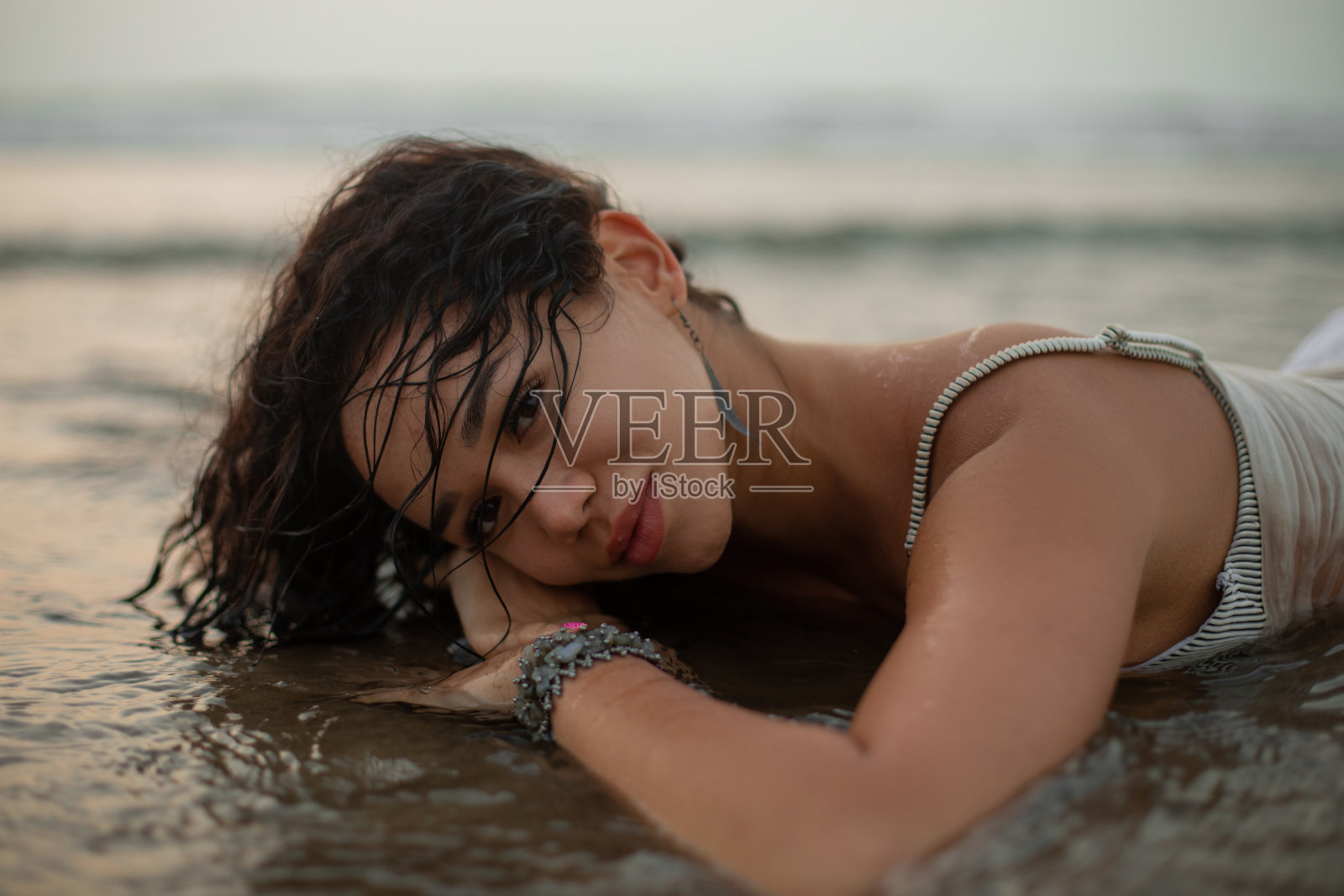 近距离特写,迷人的,微笑的卷曲的长发女人在潮湿的衣服,戴着手镯躺着,头在手在海水中照片摄影图片