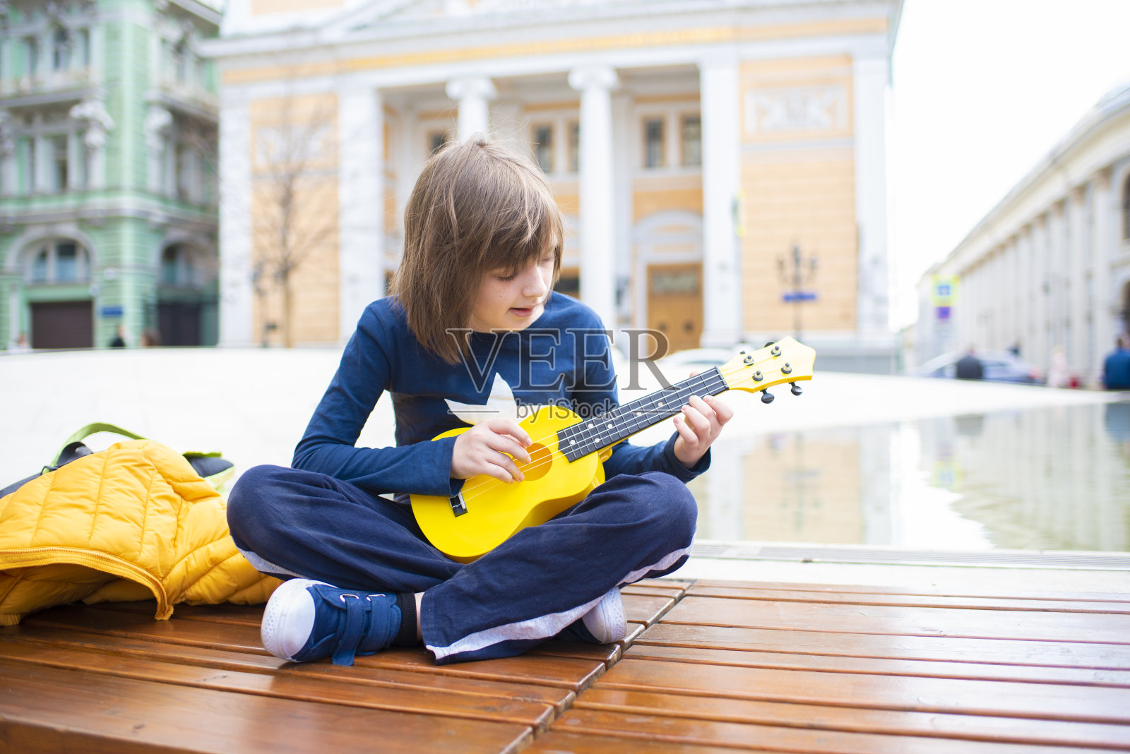 在老城的历史中心,一个十几岁的孩子坐在喷泉旁,弹着一把黄色的尤克里里琴,这是一种小吉他照片摄影图片