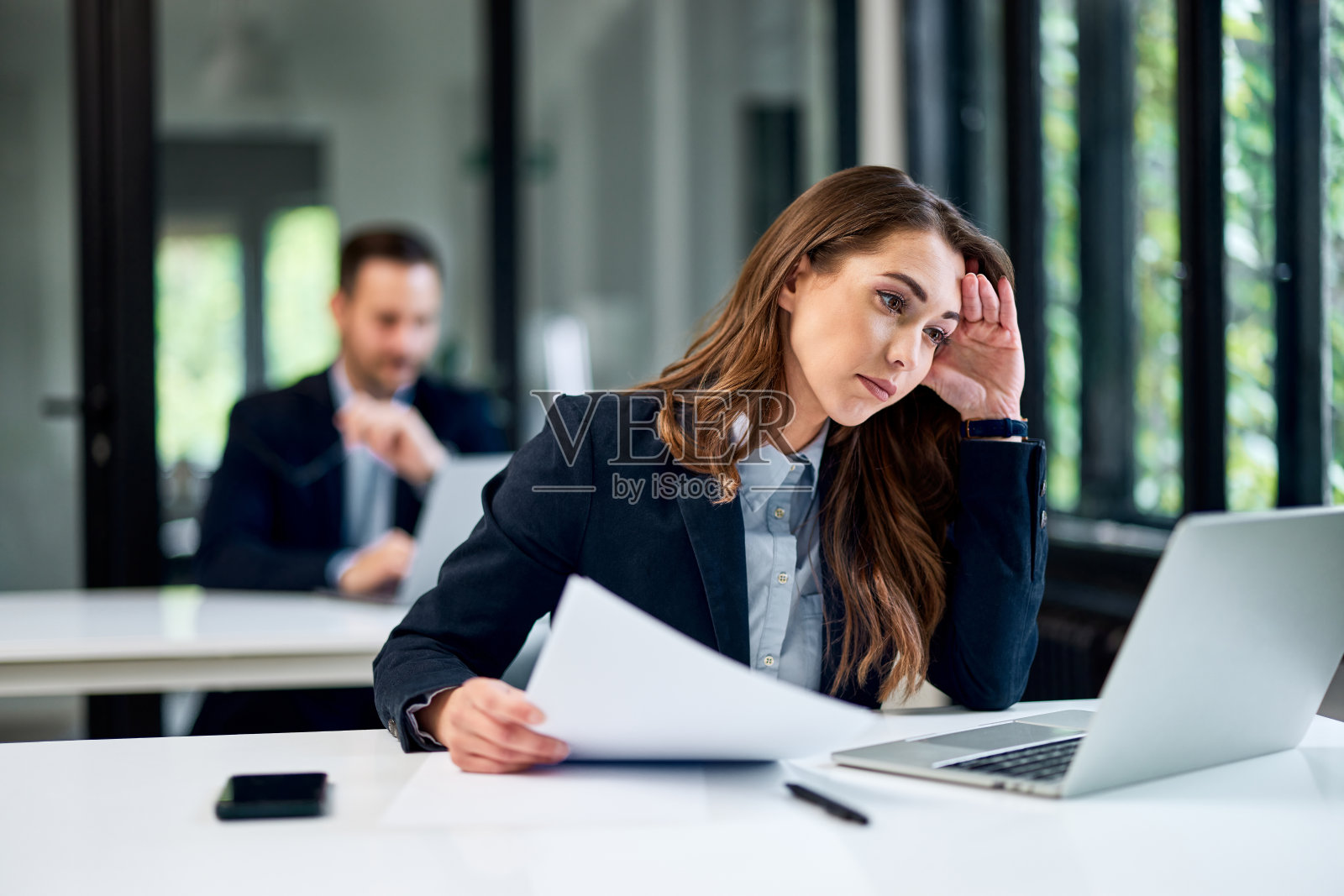 一个疲惫的女人在办公室里和一个男同事坐在一起,用笔记本电脑工作。照片摄影图片