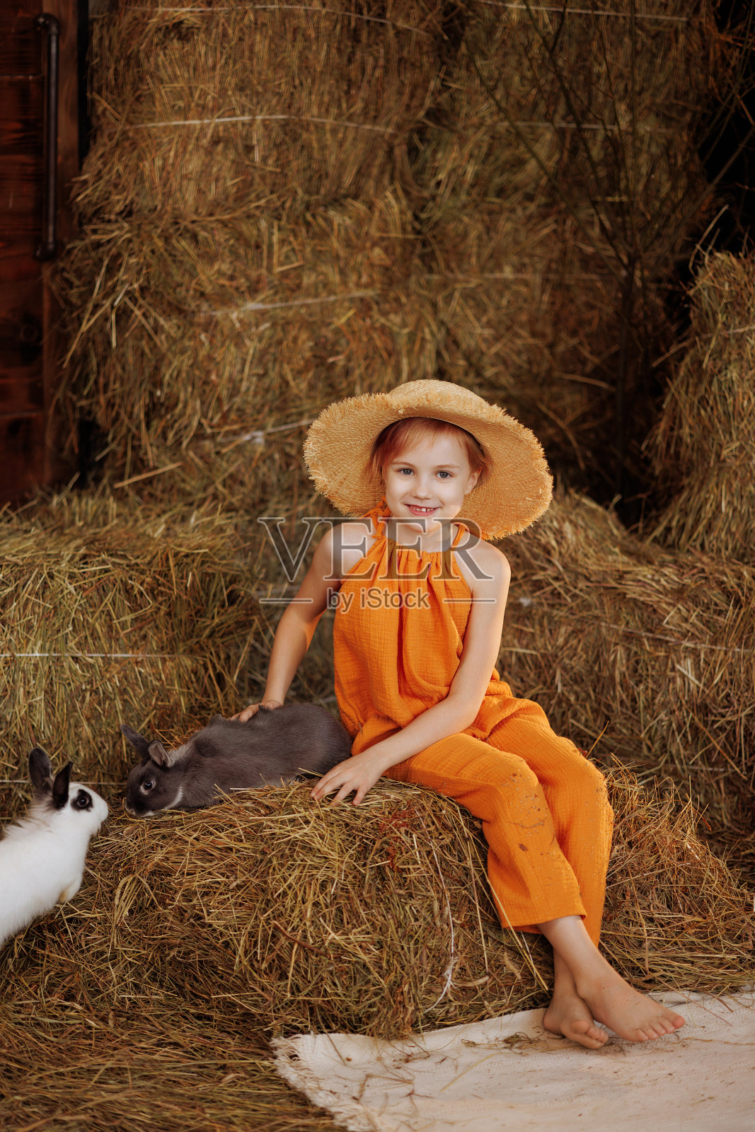 小女孩穿着橙色的裙子,戴着草帽,和一只白色和棕色的兔子坐在稻草捆上照片摄影图片