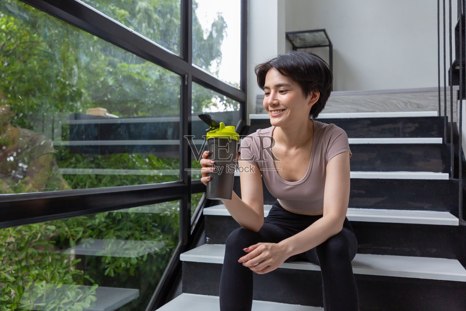 运动员女孩穿着黑色紧身运动服,拿着一个饮料瓶坐在楼梯上。照片摄影图片