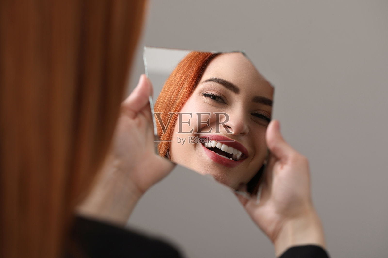 微笑的年轻女子在浅灰色背景下看着破碎镜子中的自己,特写照片摄影图片