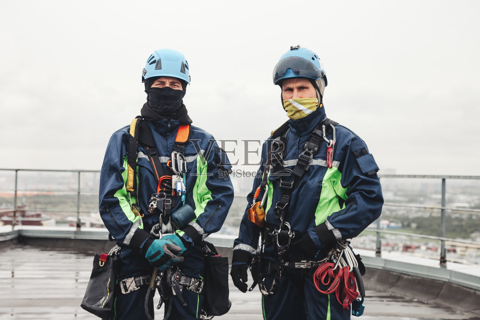 两个工业登山工人穿着制服,戴着头盔,在住宅楼的屋顶上摆姿势,看着镜头。访问工人在屋顶高层工作。工业城市工程的概念。复制广告位照片摄影图片