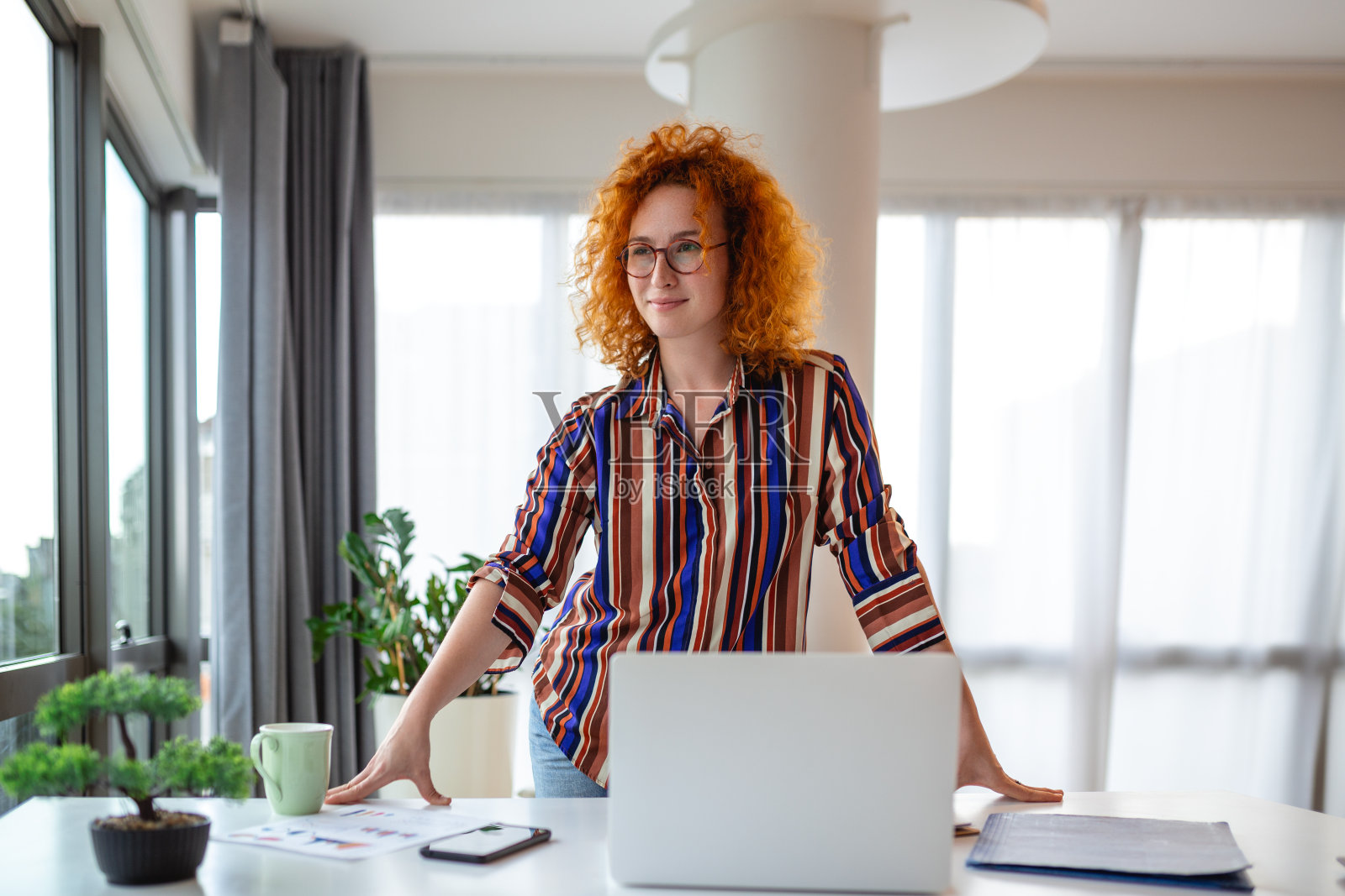年轻的女员工微笑着站在办公室的办公桌前,遥望着远方,思考着事业的成功。快乐的女商人在职场计划或梦想。商业愿景概念。照片摄影图片