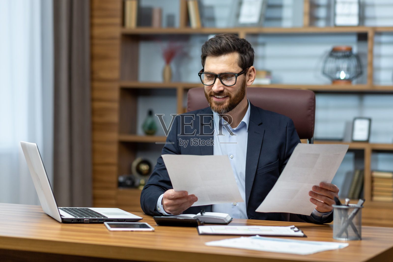 一个穿着休闲衬衫的男人对结果很满意,他手里拿着合同和报告,微笑着开心地在办公室里用笔记本电脑做文书工作照片摄影图片