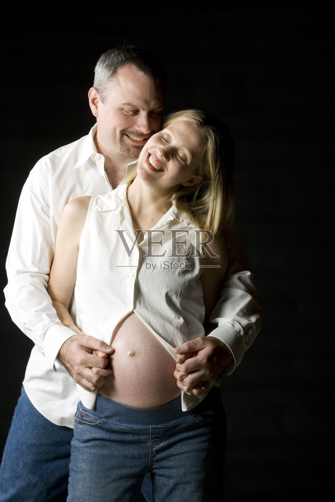 一对怀孕夫妇在黑色背景下拥抱在摄影棚里。照片摄影图片
