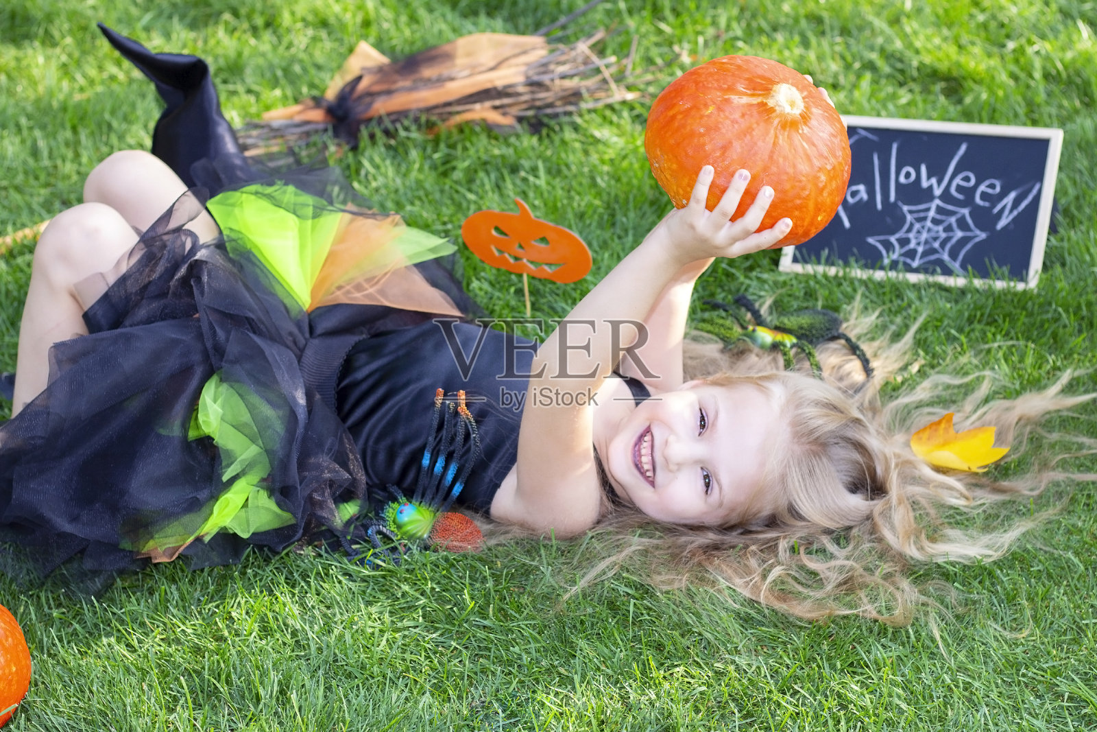万圣节穿着女巫服装的女孩。贴着标语:万圣节。快乐的女孩躺在草地上照片摄影图片