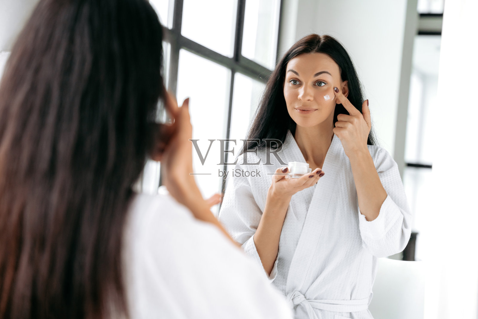 穿着白色浴袍的黑发美女站在浴室的镜子前,脸上抹着润肤霜,面带微笑。皮肤的营养和水合作用照片摄影图片