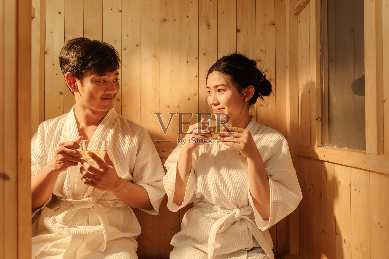 幸福的亚洲夫妇穿着浴袍喝着热茶,在桑拿浴室放松。在木制水疗中心放松的年轻夫妇。健康和水疗治疗理念。照片摄影图片