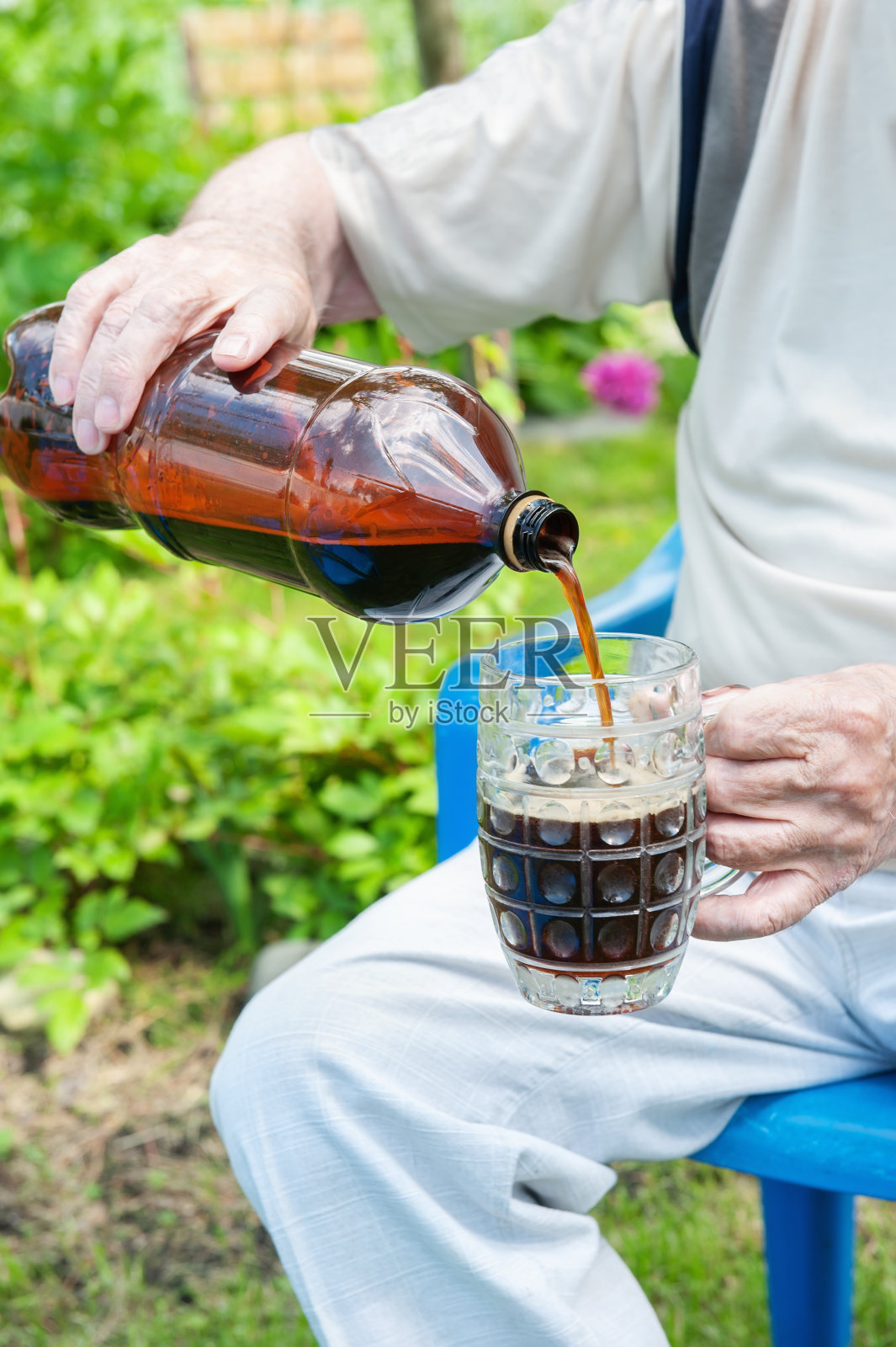 一位上了年纪的男子正用手把满是泡沫的黑色饮料从瓶子里倒进玻璃杯里。一个人在花园里干活儿后解渴。男人往杯子里倒了一杯黑啤酒照片摄影图片