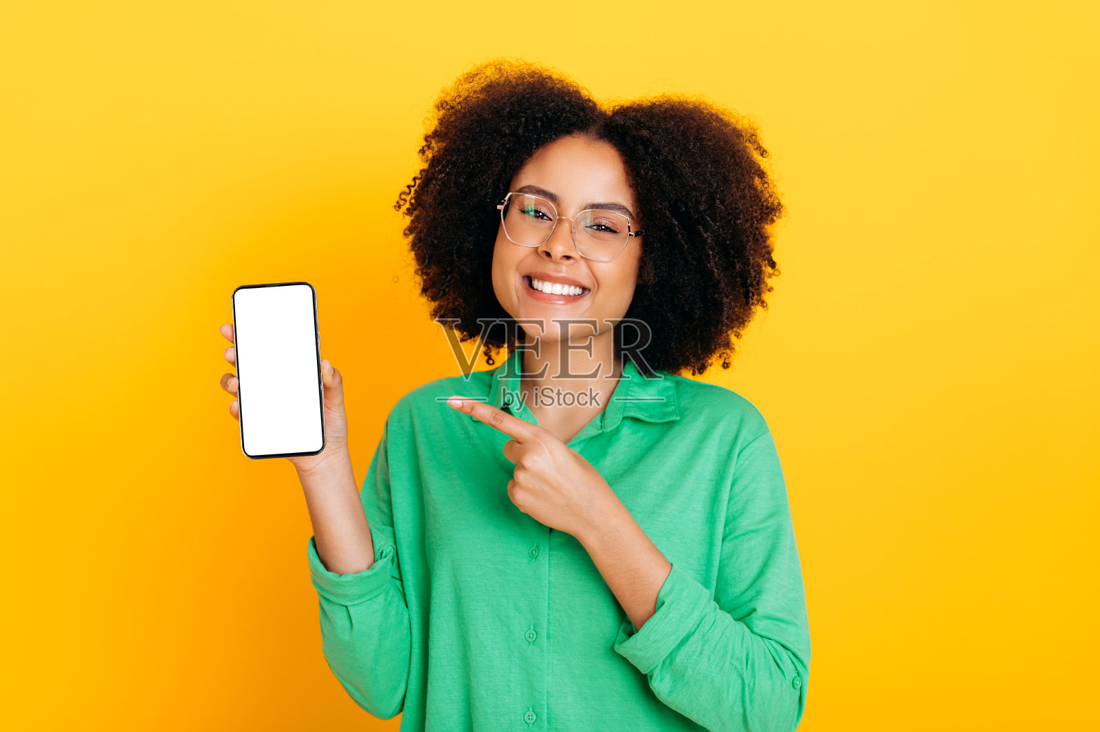 快乐的非裔美国人或巴西女人戴着眼镜,拿着智能手机,上面有空白的演示或广告模拟屏幕,指着它,对着镜头微笑,孤立的黄色背景照片摄影图片