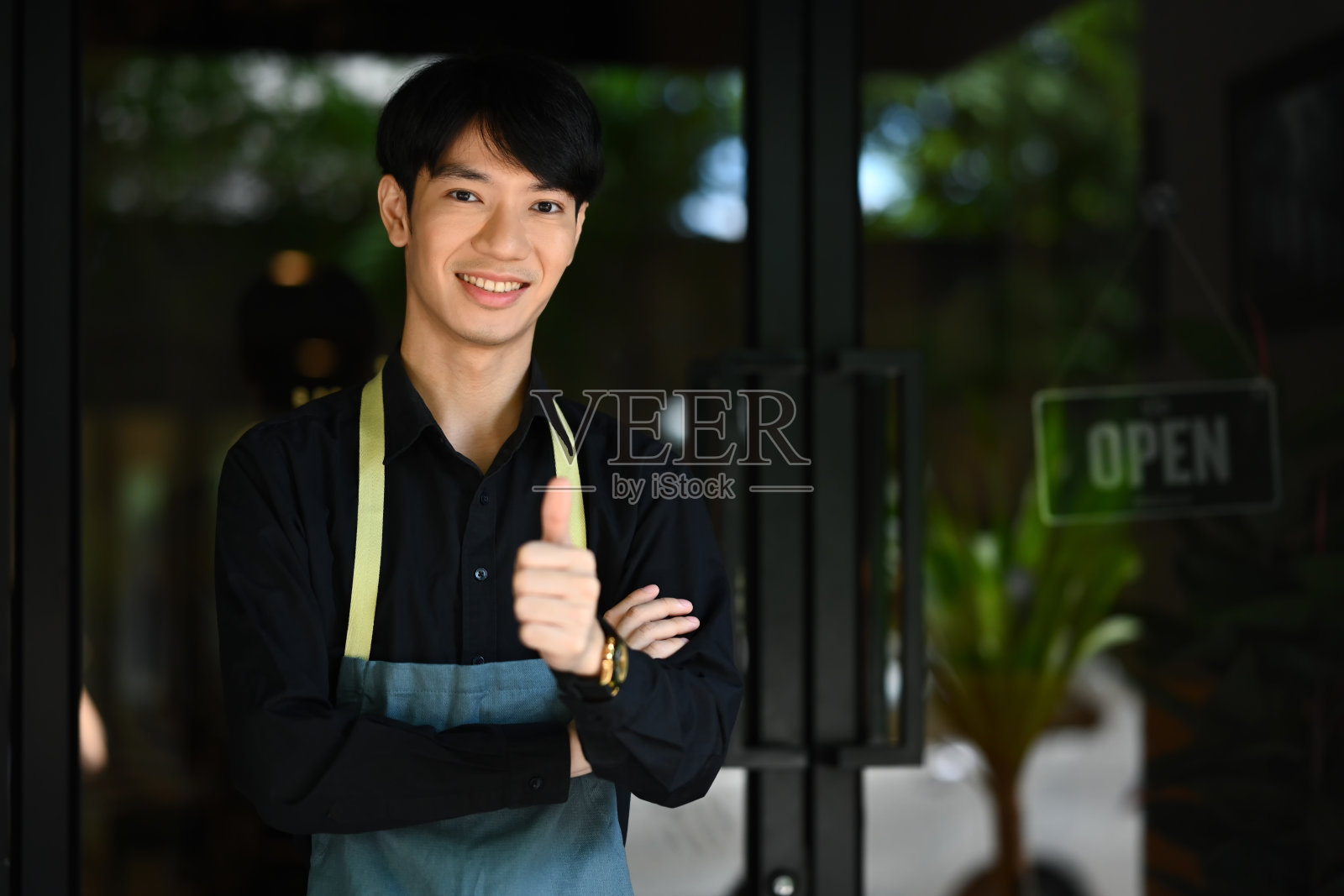 帅气的亚洲男服务员的肖像,穿着围裙,站在咖啡店前,对着镜头微笑。照片摄影图片