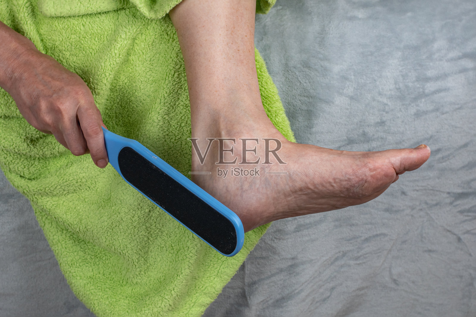 雌性用磨砂刷去脚上的角质。女性脚后跟破裂的治疗。一名妇女用塑料磨碎器对脚跟皮肤进行整容手术。愈伤组织去除概念照片摄影图片