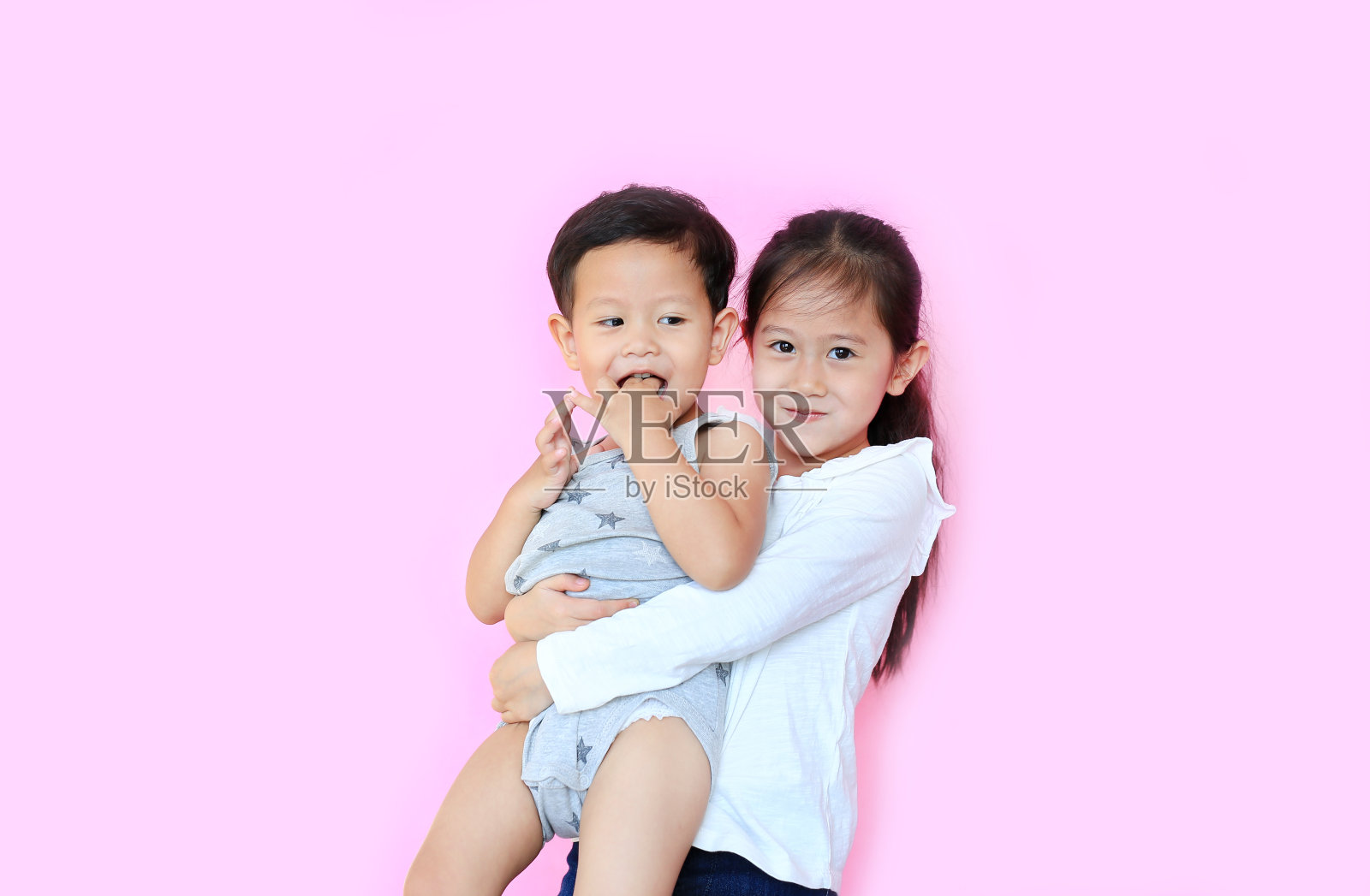 快乐的亚洲妹妹拥抱她的小弟弟孤立在粉红色的背景。小姑娘抱着她的弟弟。照片摄影图片