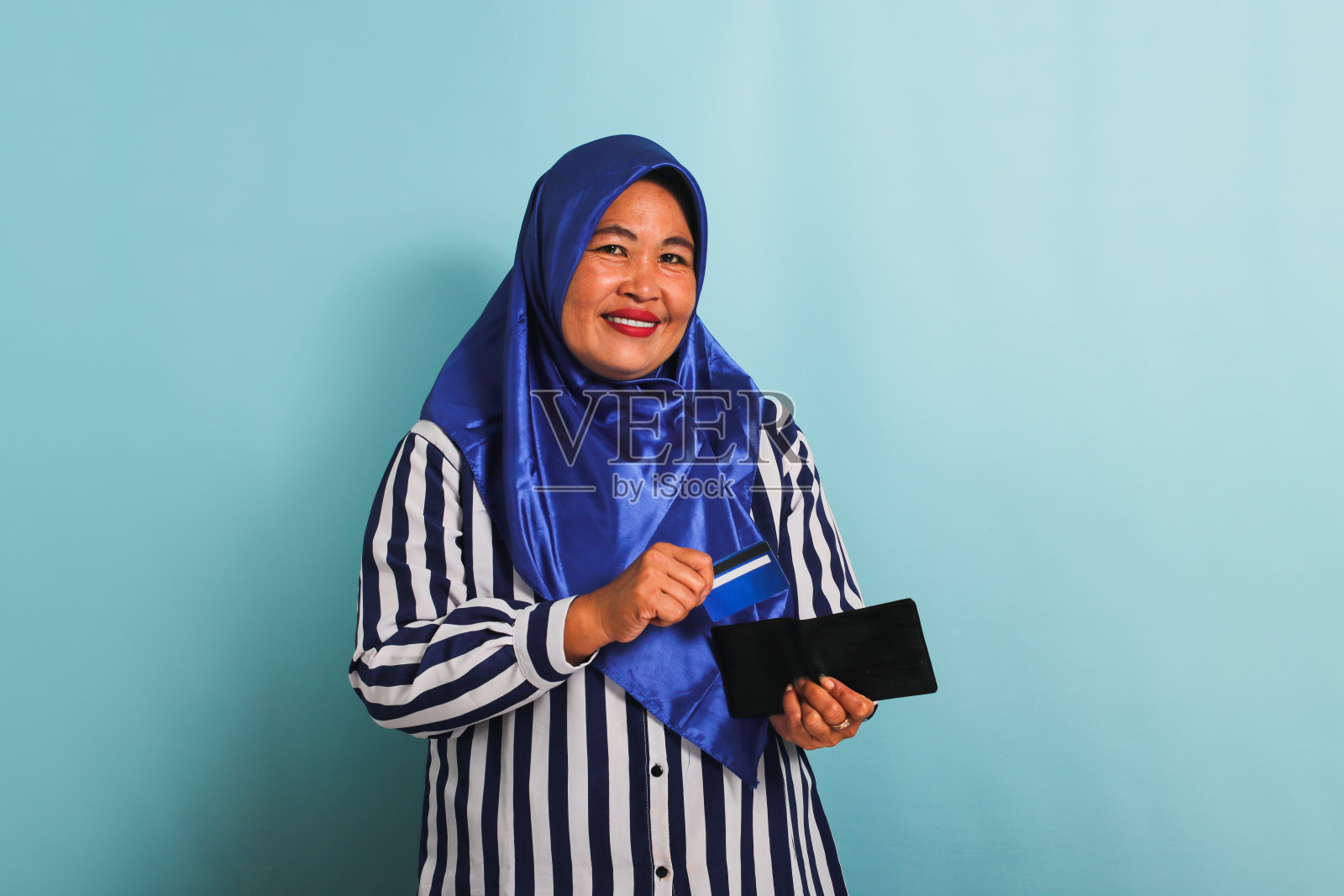 戴头巾的中年亚洲妇女从钱包里拿出信用卡,在蓝色背景中孤立照片摄影图片