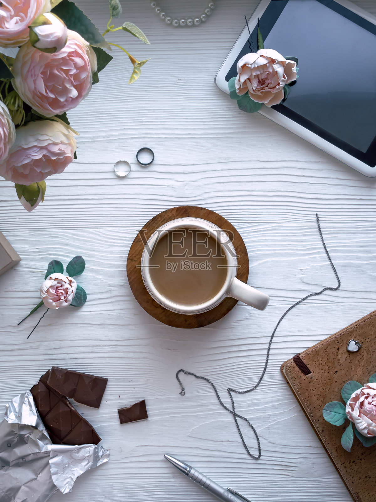 加牛奶的咖啡被倒进木桌上的杯子里。桌子上有记事本、平板电脑、钢笔、鲜花、珠宝。女性的桌面。白色背景。从高处俯瞰照片摄影图片