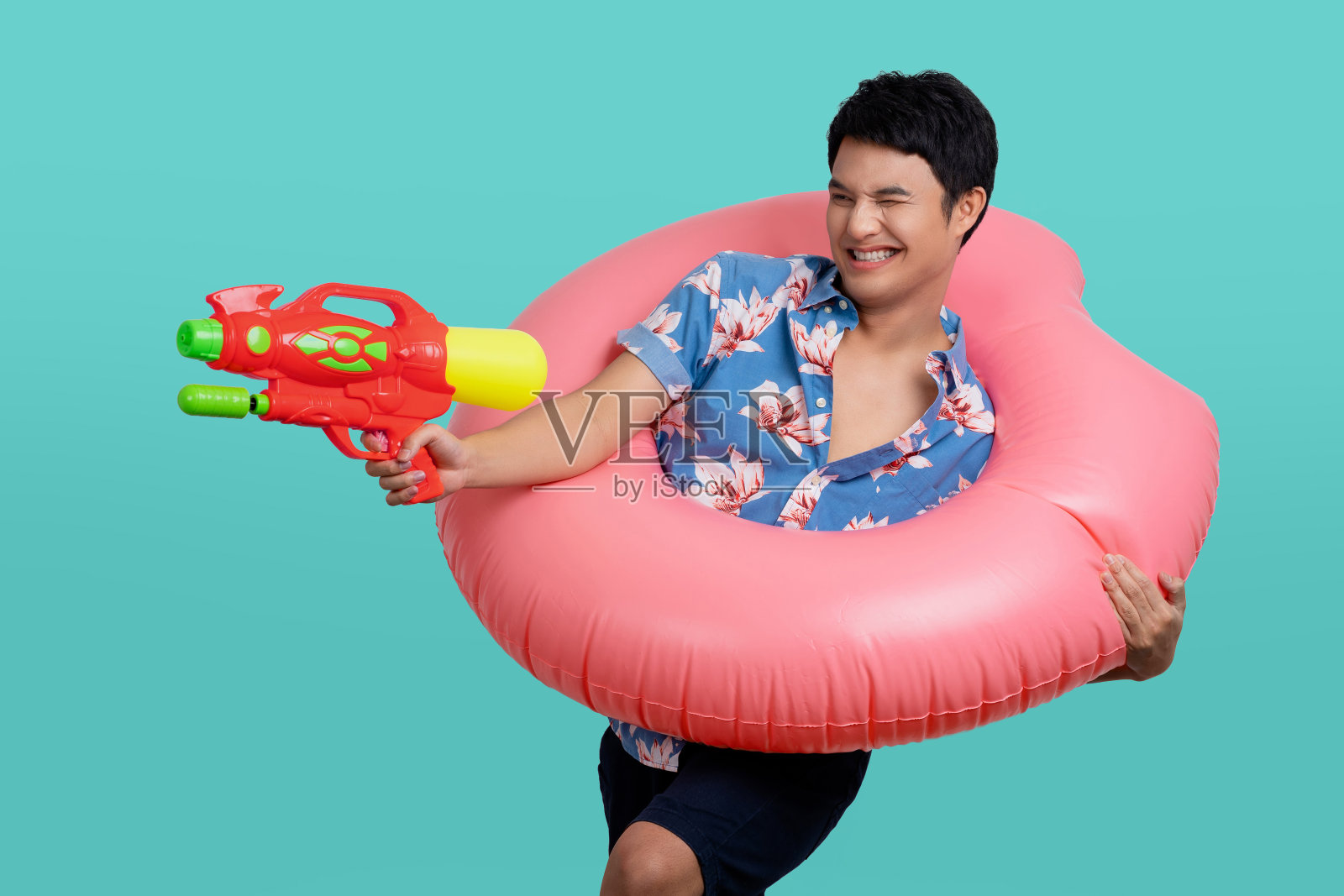 年轻的游客穿着沙滩衬衫手持充气环玩射击从水枪孤立在浅蓝色的背景。照片摄影图片