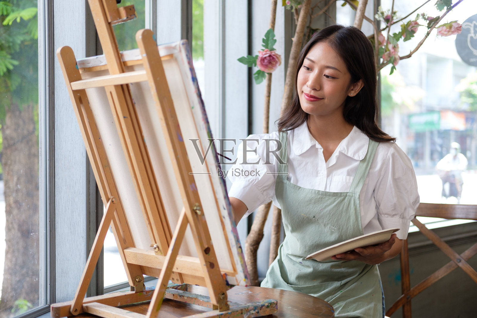 女子在画室里用油画颜料在画布上作画。女艺术家在车间画画。年轻的女艺术家在家里拿着调色板,用画笔在画布上作画照片摄影图片