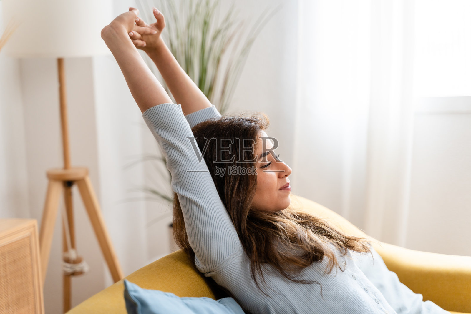 年轻女子坐在家里的沙发上伸伸胳膊。照片摄影图片