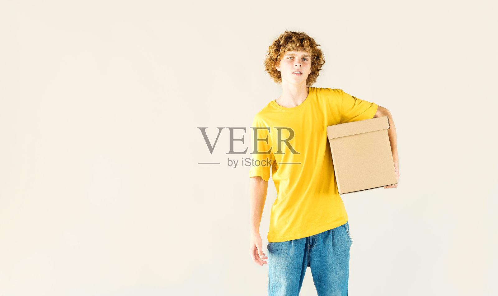 年轻卷曲的男人手里拿着包裹纸板箱的模型。带有副本空间的包裹递送模板。摄影工作室。照片摄影图片