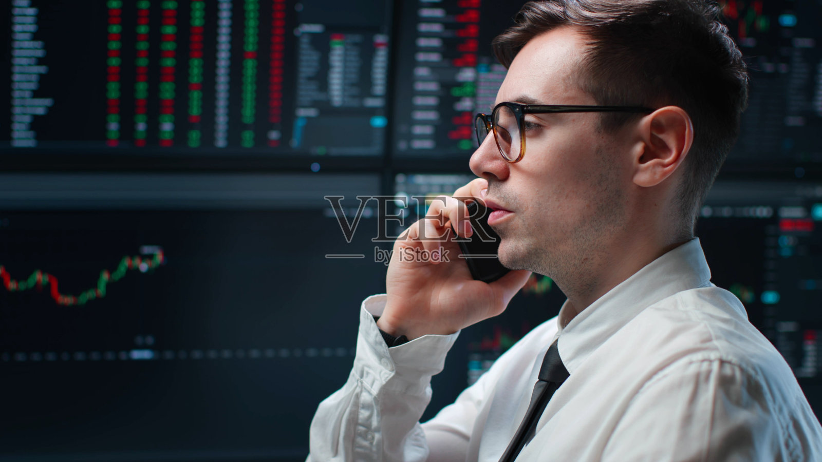 股市经纪人用手机讲话,背景是多台电脑屏幕上的图表照片摄影图片