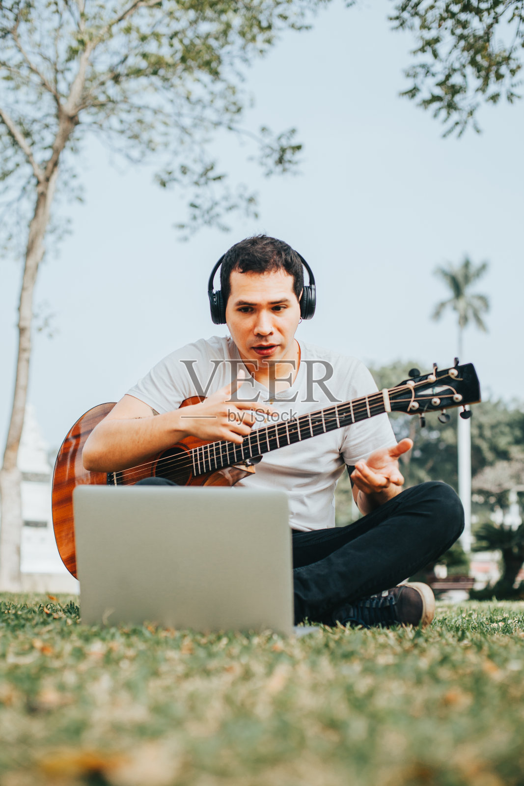 一个年轻人在公园里一边弹吉他一边用笔记本电脑教学。照片摄影图片