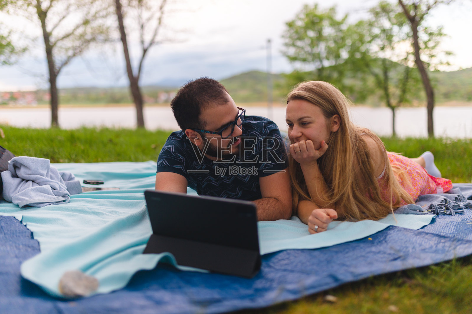 年轻浪漫的夫妇躺在野餐毯子上,在平板电脑上看电影,而在自然照片摄影图片