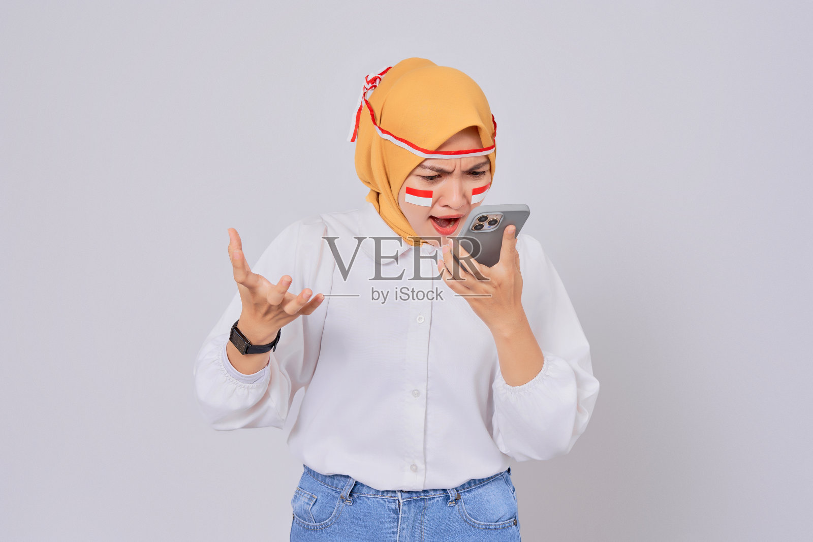 愤怒的年轻亚洲穆斯林妇女戴着头巾,对着白色背景的手机大声喊叫。8月17日庆祝印尼独立日照片摄影图片