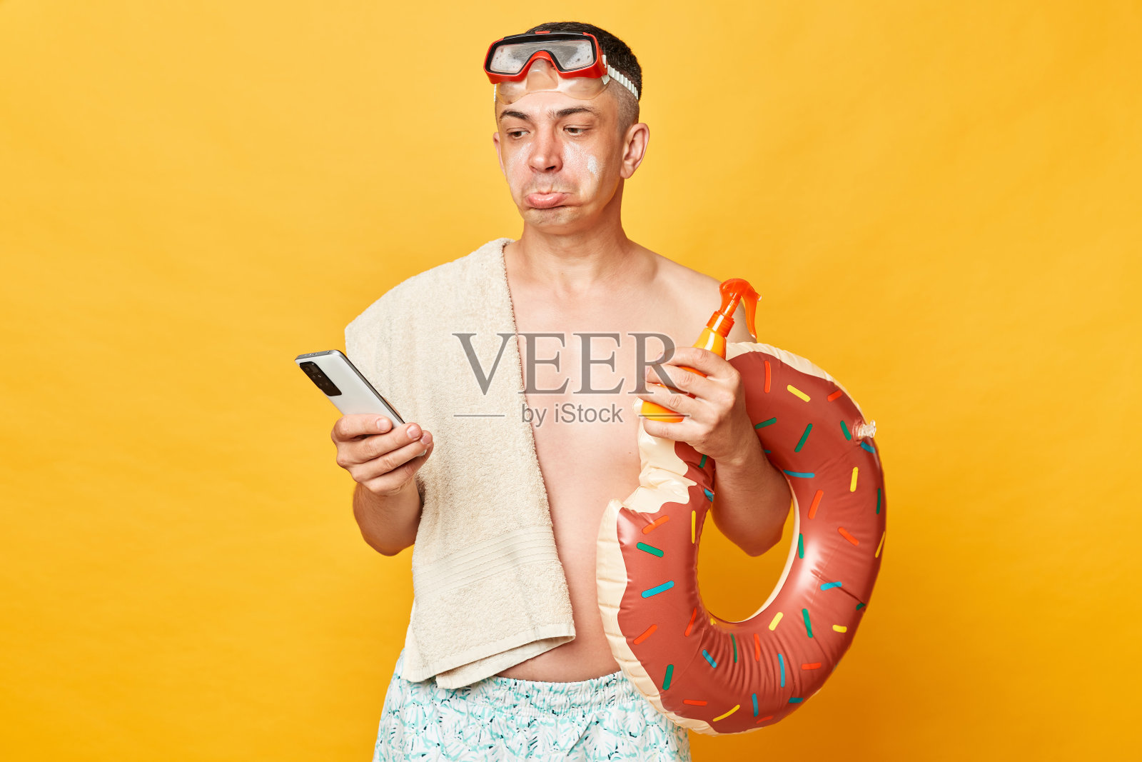 失望不高兴的成年男子穿着短裤,泳衣,潜水镜,拿着甜甜圈,橡胶圈和袋子孤立在黄色背景,拿着手机看着撅着嘴。照片摄影图片