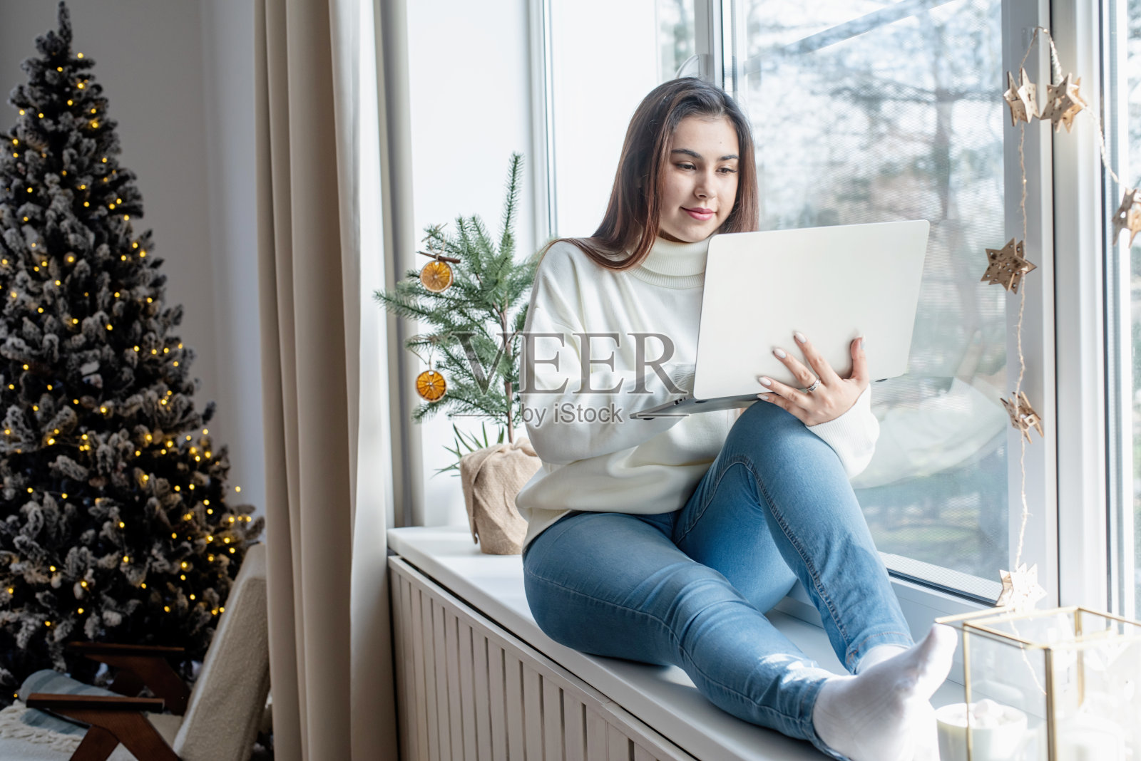 圣诞节前夕,穿着温暖的白色冬季毛衣的女人坐在家里的窗台上用笔记本电脑工作照片摄影图片