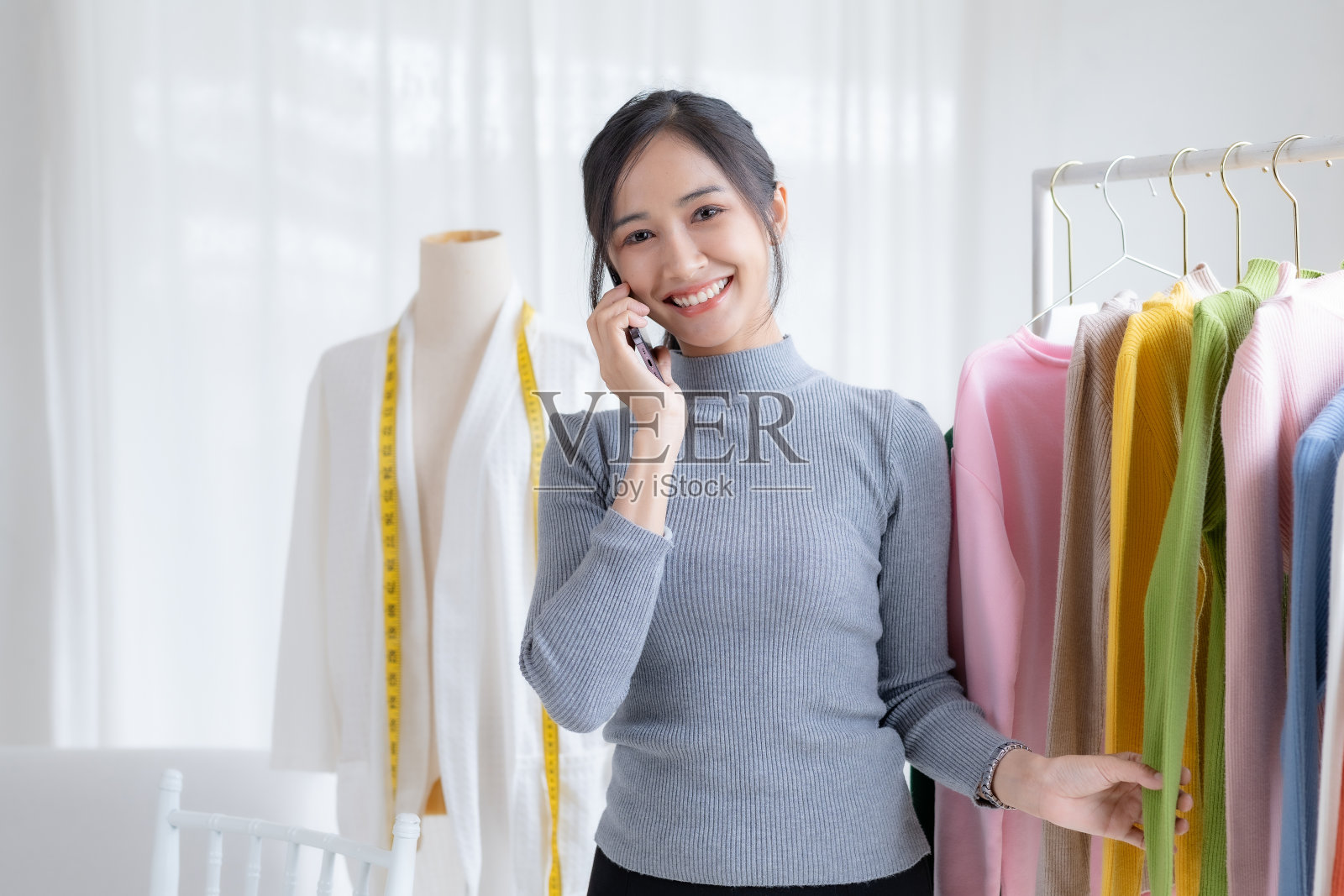 服装老板在打电话。美丽的亚洲在线企业主,他们在网上销售布料,并使用笔记本电脑解释产品的特点,价格和特点。照片摄影图片