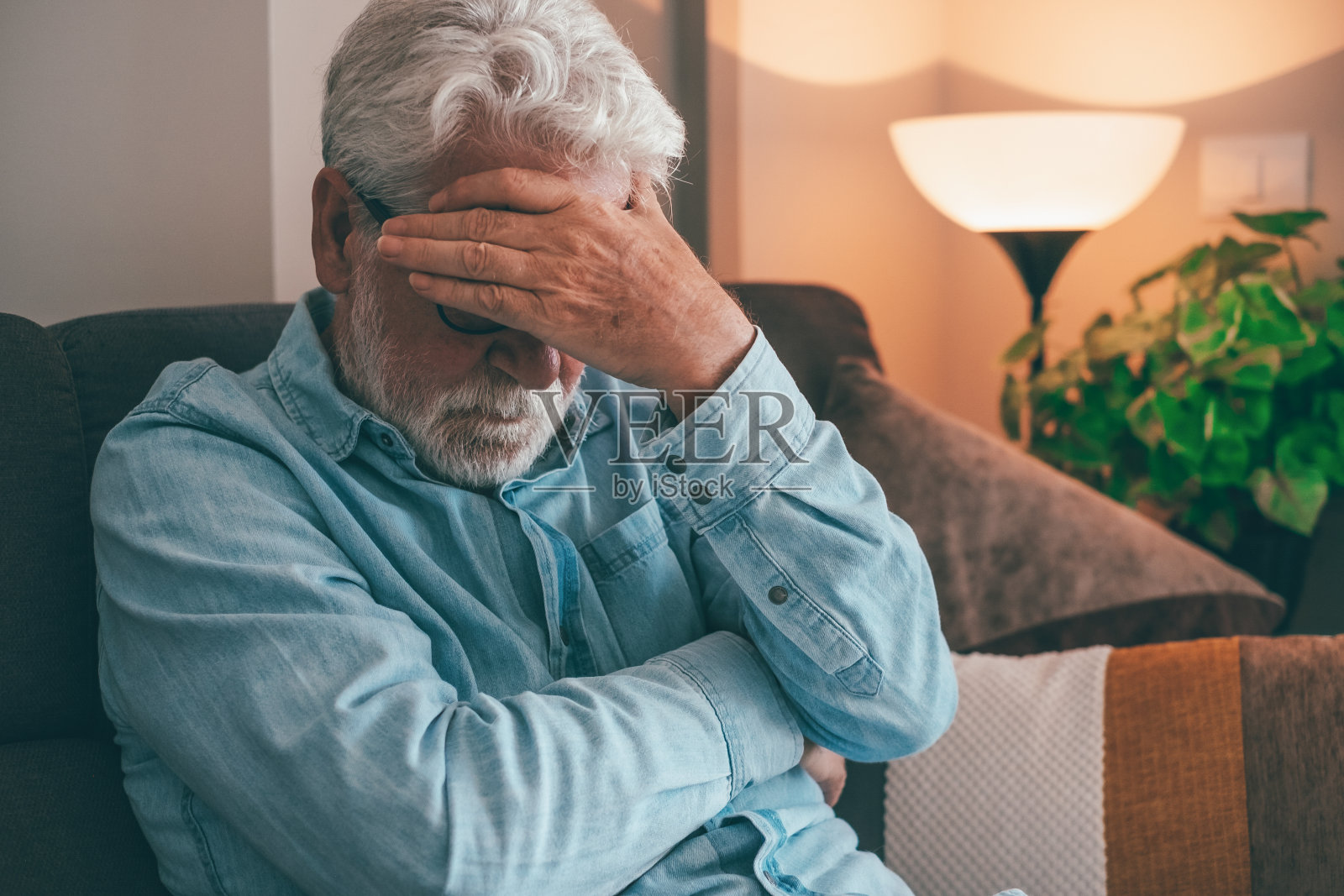 闷闷不乐的老人坐在家里的沙发上,一只手捂着脸。因经济或健康问题、坏消息、孤独、感到孤独和悲伤的退休老人照片摄影图片