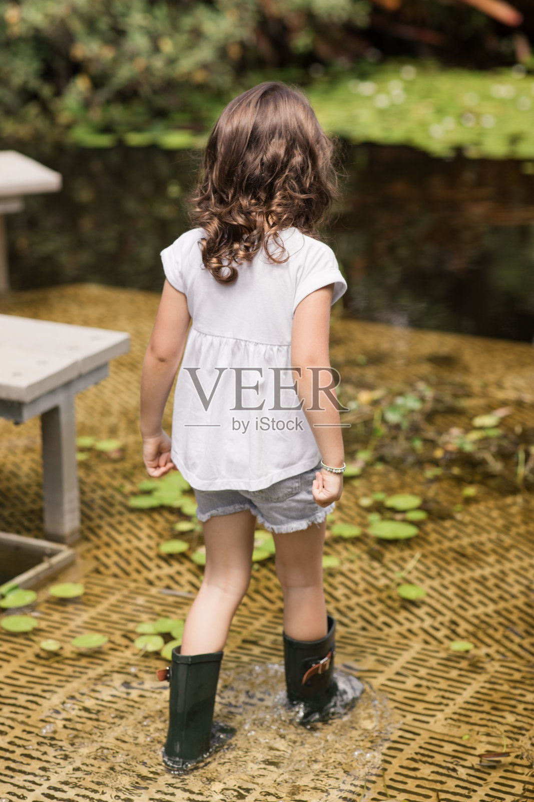 一个4岁的小女孩在炎热的夏日里穿着白色t恤,戴着Deium花,穿着深绿色雨靴,在佛罗里达州西棕榈滩花园散步照片摄影图片